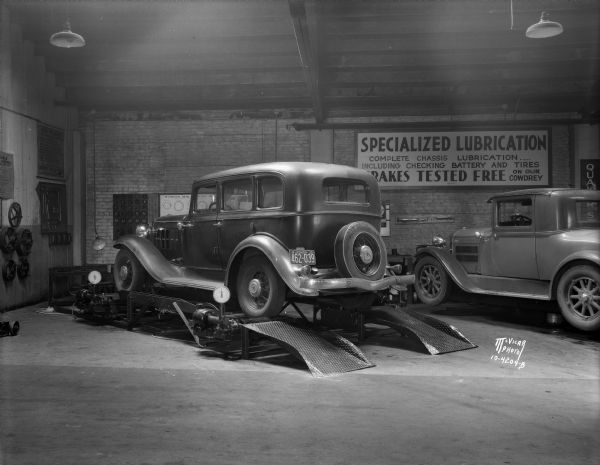 Interior view of the J.A. Brady Garage, 1307-1309 Williamson Street, owned by John A. Eichman, with a sedan on the hoist and a coupe on floor. The brakes of the sedan are being checked using a Cowdery machine, a service provided free by the garage.