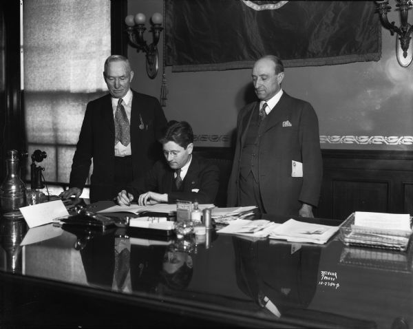 Governor Philip La Follette, the son of Senator Robert M. La Follette, Sr., signing a gas tax bill.  Senator John E. Cashman, author of the bill, stands on the left, and Assemblyman Henry Ellenbecker, a veteran Progressive, stands on right.  Until 1925 the state highway system was funded through license registrations and the property tax.  In that year a two-cent per gallon tax created the segregated highway fund.  The 1931 bill signed by Governor La Follette increased the tax to four cents per gallon.
