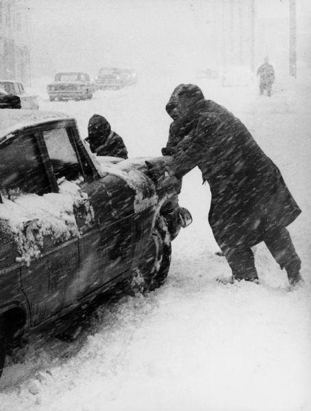 A winter scene with passers-by helping a stranded Madison motorist during a late winter blizzard.