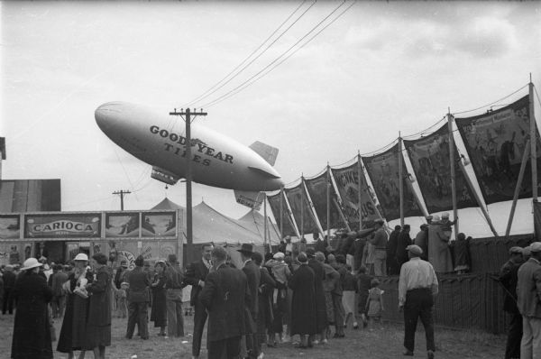 The blimp of the Goodyear Tire Company hovers above the carnival area of the Wisconsin State Fair, tethered to the ground with ropes.  The fairgoers, however, seem uninterested.
