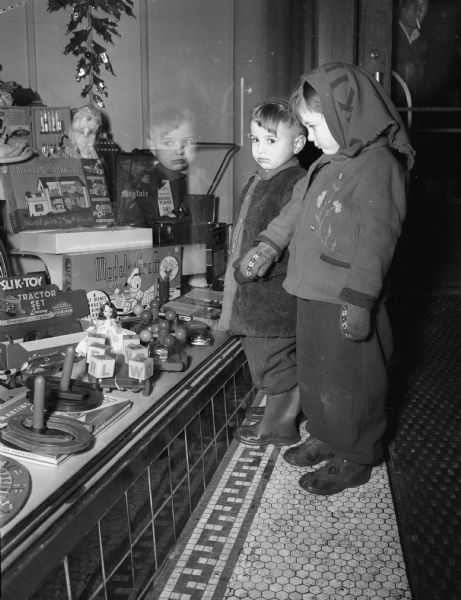Two children, a boy and a girl, look at display of toys in a store window.