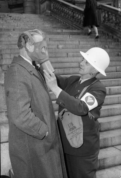 Dane County chief air raid warden, Coroner E.A. "Ace" Fischer puts a gas mask on Deputy Sheriff Albert Dahle on a stairway at the Wisconsin State Capitol.