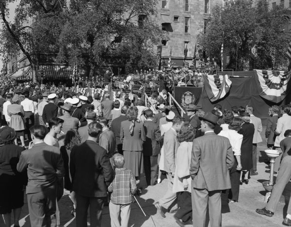 A large crowd gathered around a bandstand on Capitol Square watching a band playing at the Wisconsin State Centennial celebration.