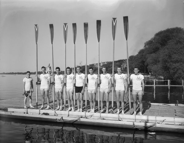 Group portrait of the University of Wisconsin eight-man crew standing on the crew pier on Lake Mendota holding their oars.
