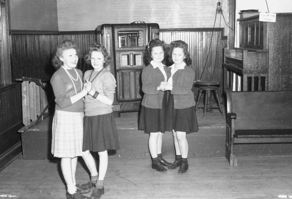 Two sets of twins dance at the recreation center for school pupils at the Cottage Grove Town Hall. Included on the left are Jean and Joan Hanson of Cottage Grove, and on the right Barbara and Betty Gunderson of Deerfield. There is a jukebox in the background, and a table along the wall on the right.