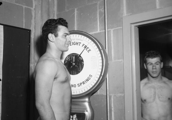A football player stands on scale weighing in at 183 lbs.  A second player is standing nearby.