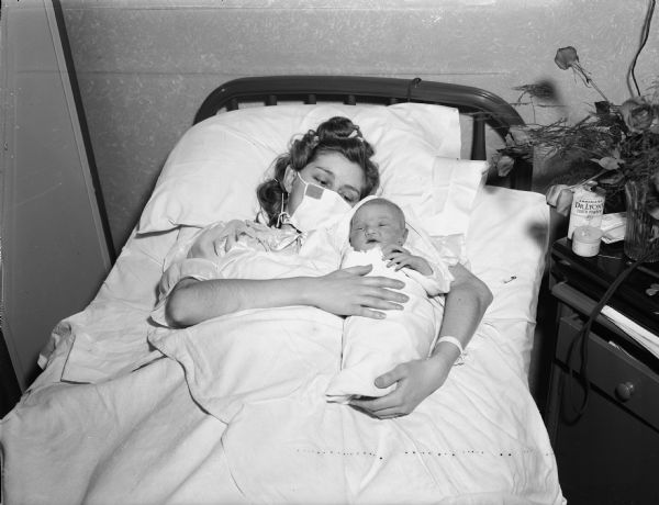 A mother, with a mask over her mouth and nose, lying in a bed holding an infant. This is probably the first baby born in Madison in 1952, just 5 seconds after midnight to Mr. & Mrs. Joseph Temkin.