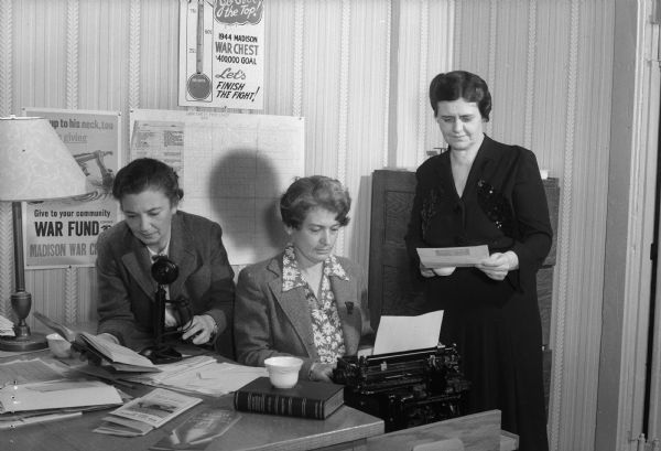 Three women work in the Community War Chest office. One of the women is typing and one is about to use the telephone. A sign on the wall behind the women reads: "1944 Madison War Chest $40,000 goal, Let's Finish the Fight!" Madison War Chest contributes money to the National War fund to support 21 voluntary agencies working for GIs still in service, including USO, and the Allies.