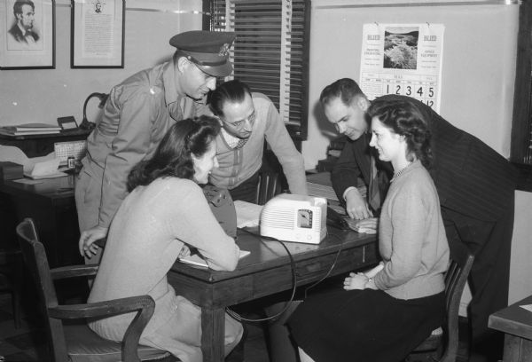Two women and three men (one in uniform) gathered around a table listening to a radio commentary on V-E (Victory in Europe) Day.