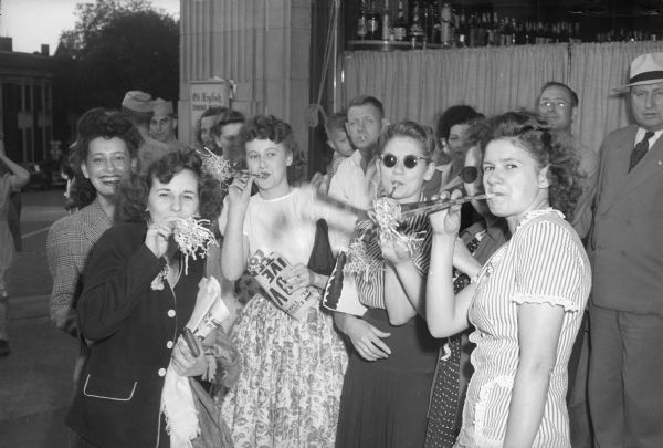 Four young women blowing on noise makers, with other people watching, celebrating V-J Day, August 15, 1945, the day on which the Allies announced the surrender of Japanese forces during World War II. They are standing in front of a building at a street corner. The sign near the window reads: "Old English Dining Room."