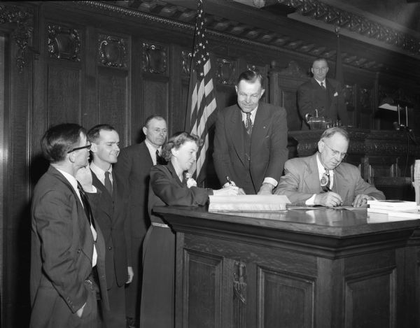 Newly-elected members of the Wisconsin Assembly signing in. Facing the camera left to right are: E. William Proxmire (D-Madison), Hermann Eisner (D-Cross Plains), and Ruth Doyle (D-Madison). Assembly Clerk Robert Boysen looks on as Mrs. Doyle signs. Seated at the deak is George Rude, an Assembly clerk. Chief Clerk Arthur L. May is behind the speaker's rostrum.  At the extreme left is Assemblyman Elmer Genzmer (R. Mayville).