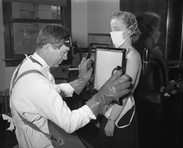 Doris (Mrs. Edward) Trameri being x-rayed by Dr. James M. Wilkie for tuberculosis at the free chest clinic operated by the Madison Department of Public Health. The doctor is wearing goggles on his head, and protective gloves. Doris is wearing a face mask.