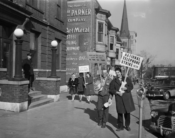 Telephone workers picket at the telephone exchange on Capitol Square, near the corner of Main and Carroll Streets. Seven hundred Madison telephone employees participated in the country's first nationwide telephone strike. Picket signs include "We'll walk day and nite until MA BELL is not so tight".