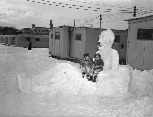 Winter scene with two young girls, Jo Ann Simon and Barbara Gatanowicz, sitting on a reclining snowman in front of the Camp Randall married student trailers at the corner of University Avenue and Breese Terrace.