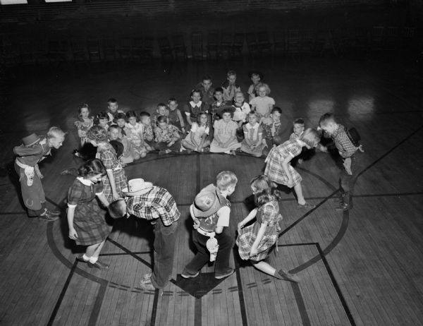 Third graders at Lincoln School as Bar-None ranch hands singing and square dancing around a "campfire" in the gymnasium. Four couples, boys dressed as cowboys and girls in plaid dresses, are bowing to one another. The rest of the children are performing as a rhythm band. The couples are (left to right): Elliott Zimmerman, Zona Sechrest, Kay Tantillo, Billy Newkirk, John Livesey, Nancy Morrison, Anthea Roberts, and Dick Lee.