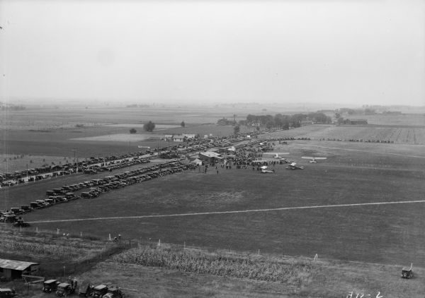 Aerial view of airplane races at the Janesville airport, including airplanes on the landing field, airport structures, cars parked around the airport, and spectators looking at the planes.