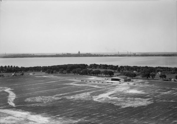Aerial view of the Pennco Field (Royal Airport), including the hanger and airplanes on the ground. Lake Monona, the Wisconsin State Capitol, and the Madison city skyline are in the background.
