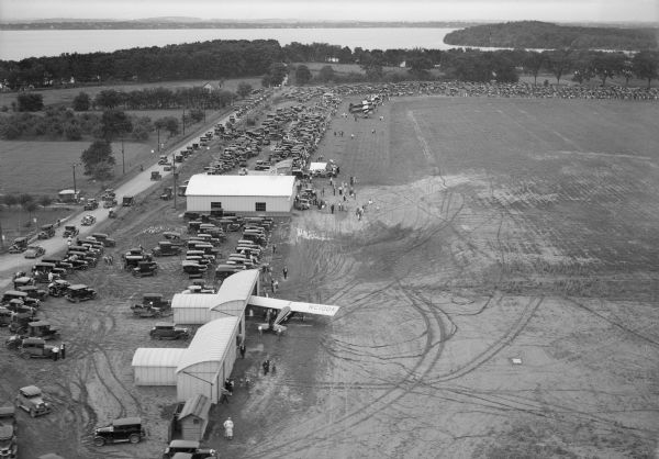 Aerial view of a crowd at the Pennco Field (Royal Airport), gathered for an aerial show.  Biplanes possibly used for the show are on the airstrip.  Automobiles are parked on the airport grounds.  Lake Monona is in the background.