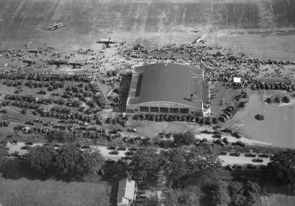 Aerial view of the Pennco Field (Royal Airport) hanger surrounded by spectators gathered for an air show.  Biplanes are parked on the grounds of the airport.  Automobiles are parked around the airport grounds.