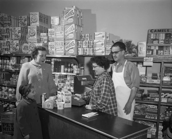 Two clerks at a grocery store checkout counter with a woman and child purchasing milk and bread. The shelves behind the register are stocked with a selection of grocery merchandise. Possibly Wilcox Food Shop, Burton & Loretta Wilcox, 1302 Mound Street.