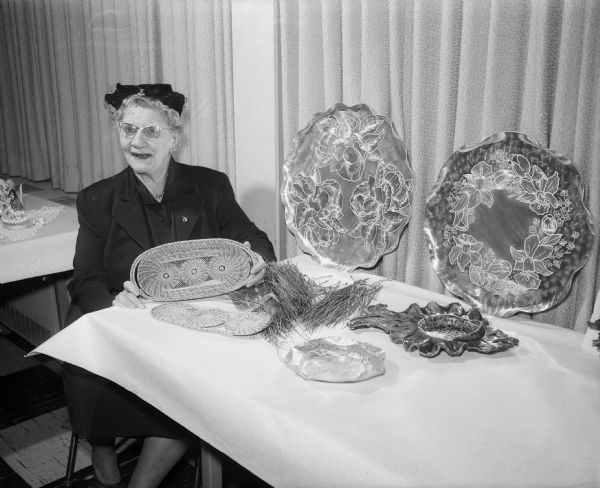 Mayme Peterson (Mrs. Alf), seated at table at East Side Women's Club hobby show with her handicrafts, including a pine needle basket.