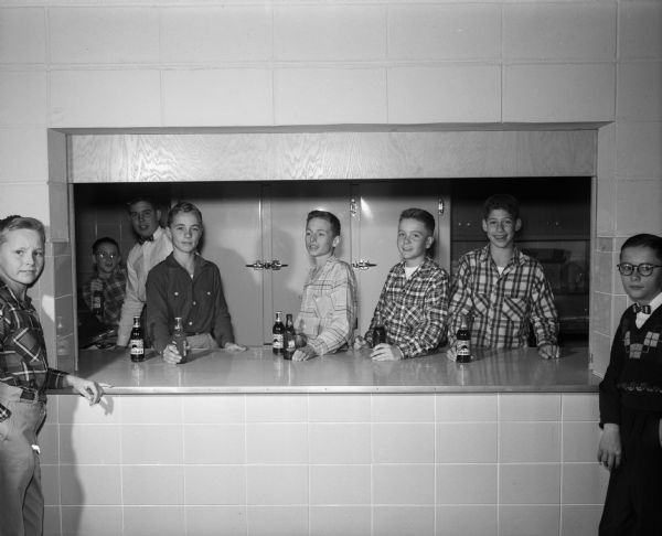 Seven boys and one teacher from Cherokee Heights School at the refreshment counter at a school dance. Four of the boys, Jerry Meulenberg, Douglas Green, Mike Weger, and Stanley Jackson, are standing behind the counter selling soft drinks.