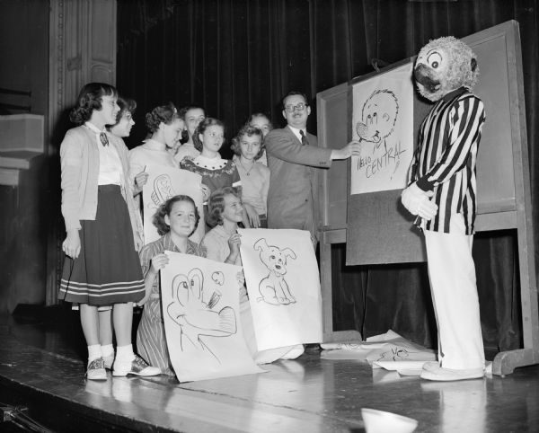 Cartoonist Walt Kelly at Central High School with a person dressed as one of his characters, Pogo. He is showing a drawing to a group of students.
