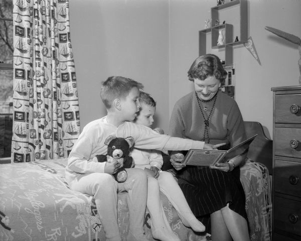 Betty J. DiSalvo reading good night stories to her children, Michael DiSalvo (age 6) and Mark DiSalvo (age 4) after having solicited money for 27 Red Feather services of the United Community Chest. The children are in pajamas and Michael is holding a teddy bear.