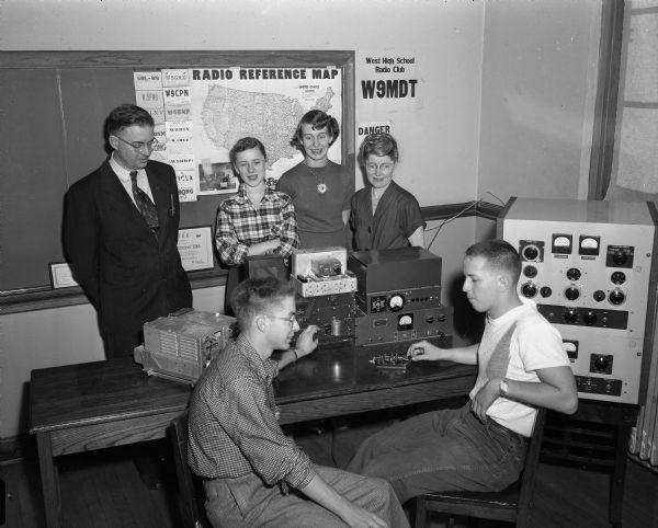 Two members of the West High School radio club, Alan Frees and Bill Reeve, operating the club's "ham" station.  Observing the young men are Henry Lugg, sponsors of the club at East High, Margaret Kincaid, Nancy Meinke, and Mrs. Lugg, adviser of the West High club.