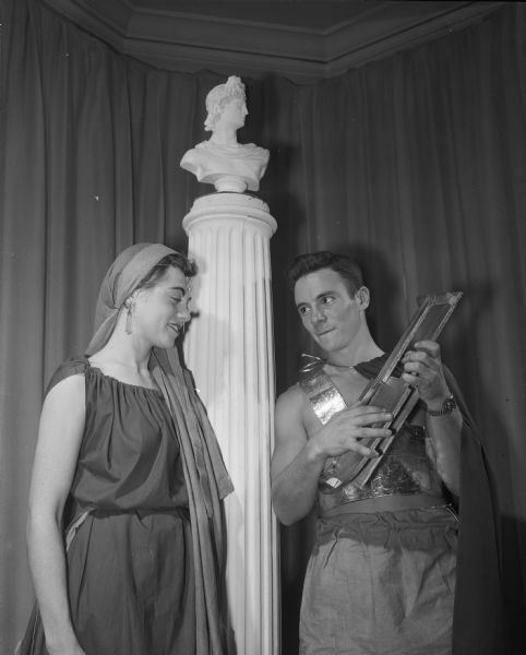 Phi Kappa Fraternity member strumming a lyre and entertaining a female guest, both in Greek costumes, at a Toga Party Dinner Dance.