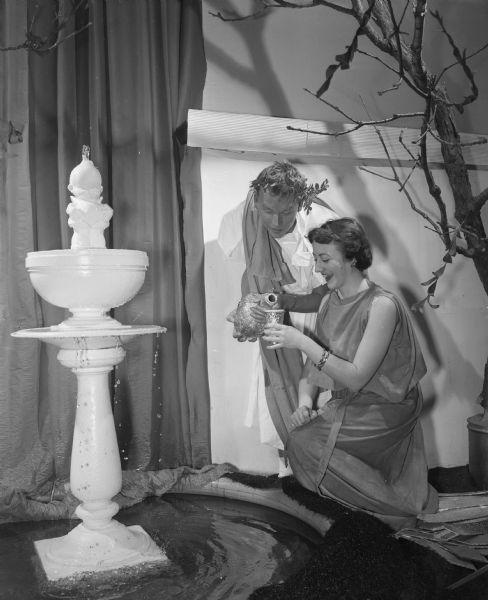 Two guests at the Phi Kappa Fraternity Toga Party Dinner Dance drinking by a Grecian fountain.