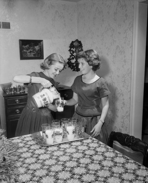 Two West Junior High School eighth graders, Patty Radder and Jean Kelzenberg, pouring glasses of milk for their potluck club supper held at the Radder home on the occasion of Patty's 13th birthday.