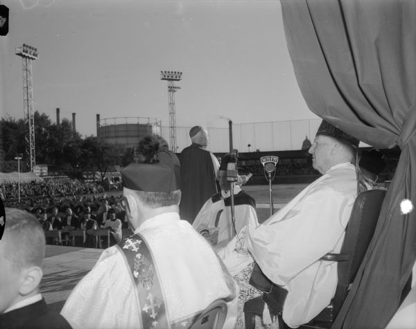 Archbishop Philip Pocock of Winnipeg preaching the sermon at the Confraternity of Christian Doctrine pontifical field mass held at Breese Stevens Field.  The Most Reverend John Treacy, bishop of the La Crosse Diocese and celebrant of the pontifical mass, is seated in the foreground wearing the mitre.