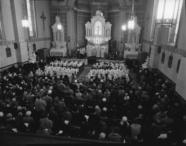 Historic evening mass at St. Raphael Cathedral, a stately two-hour ceremony closing the parish's annual 40 Hours' Devotion. This was the first evening mass held in Wisconsin and possibly the nation after authorization from Pope Pius and Bishop William P. O'Conner. Interior view of the church including the altar with the celebrants and the overflowing crowd.