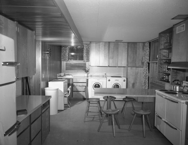 A modern kitchen including a breakfast bar with four three legged stools and a youth chair, and the laundry room with the washer, dryer and mangle.
