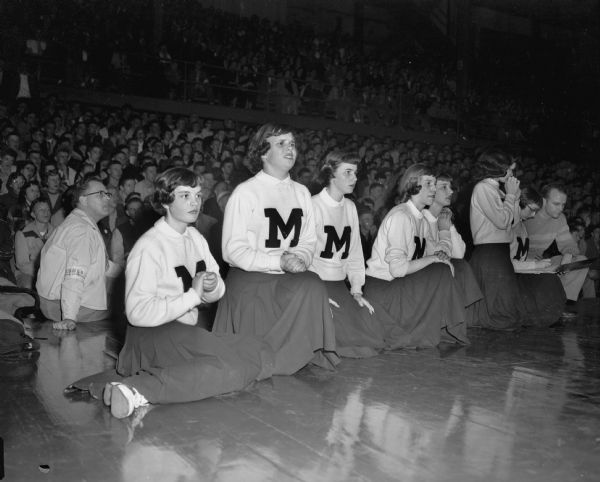 Seven female cheerleaders from Menasha High School watching a basketball game at the WIAA State Basketball Tournament.