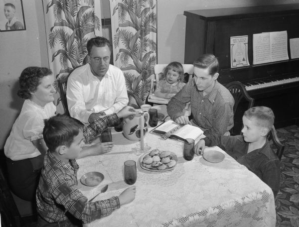 A family sits around a table eating a snack and listening to the older son reading from the bible.