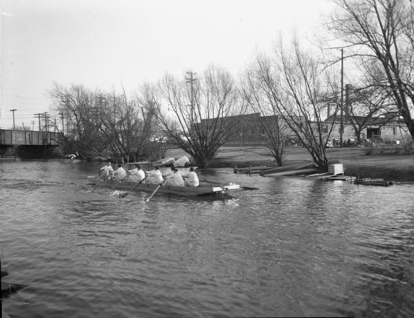 University of Wisconsin varsity crew in a practice boat on the Yahara River.  View looking south with Westinghouse Electric Supply Company, 110 North Thornton Avenue, in background.