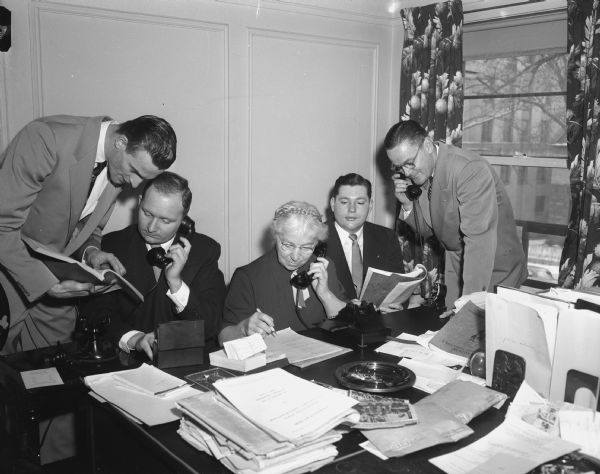 Four men and one woman seated around a table make telephone calls.