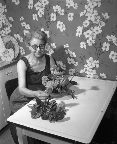 Glenn M. Wise (Mrs. John Wise), Wisconsin's first woman Secretary of State, arranges a floral centerpiece on a table in front of a floral pattern of wallpaper.