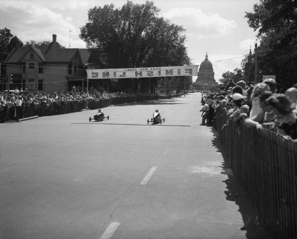 The Soap Box Derby held on East Washington Avenue leading from the Wisconsin State Capitol. Shown are two of the more than 100 racers crossing the finish line with the crowd along the course and the Wisconsin State Capitol building in the background.