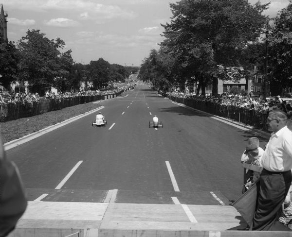 The Soap Box Derby held on East Washington Avenue leading from the Wisconsin State Capitol. Shown are two racers heading down the course from the starting ramp with the crowd gathered along the course.