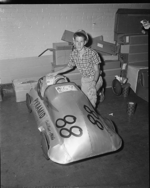 A young man poses with his Soap Box Derby car indoors. The Derby was held on West Washington Avenue leading from the Wisconsin State Capitol.