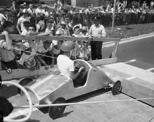 A contestant in Madison's Soap Box Derby in his racer on the starting ramp with crowd in the background. The Derby was held on East Washington Avenue leading from the Wisconsin State Capitol.