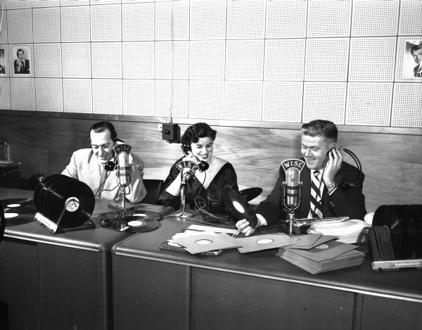 Ralph O. Connor, manager of WISC; Carol Cowan, known professionally as "Barbara Lane," and Henry Buslee, a staff member at WISC, sitting at a table behind microphones, participating in an Empty Stocking club radio marathon.
