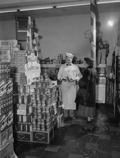 A woman purchases a can of Del Monte pineapple from a male clerk at Hi-Lo Supermarket, 2038 Jenifer Street. They are standing in front of a Del Monte "Sunshine Cruise" display.