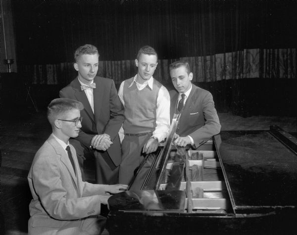 Three young men watch another young man play the piano.