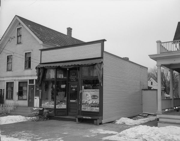 Exterior view of Harry's Food Shop, 1122 Chandler Street, owned by Harry and Loretta Blodau. A baby buggy and a doll stroller are parked outside on the sidewalk. A home is next door.