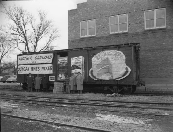 Six people are standing in front of a railroad car with a Duncan Hines advertisement on the side, two boxes of cake mix and a large cake with the sign: "Another Carload of Duncan Hines Mixes," sponsored by Certified Foods.