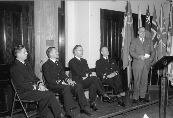 Five men on a stage at a vocational school.  Four of the men are in military uniforms, while one is dressed in a business suit.